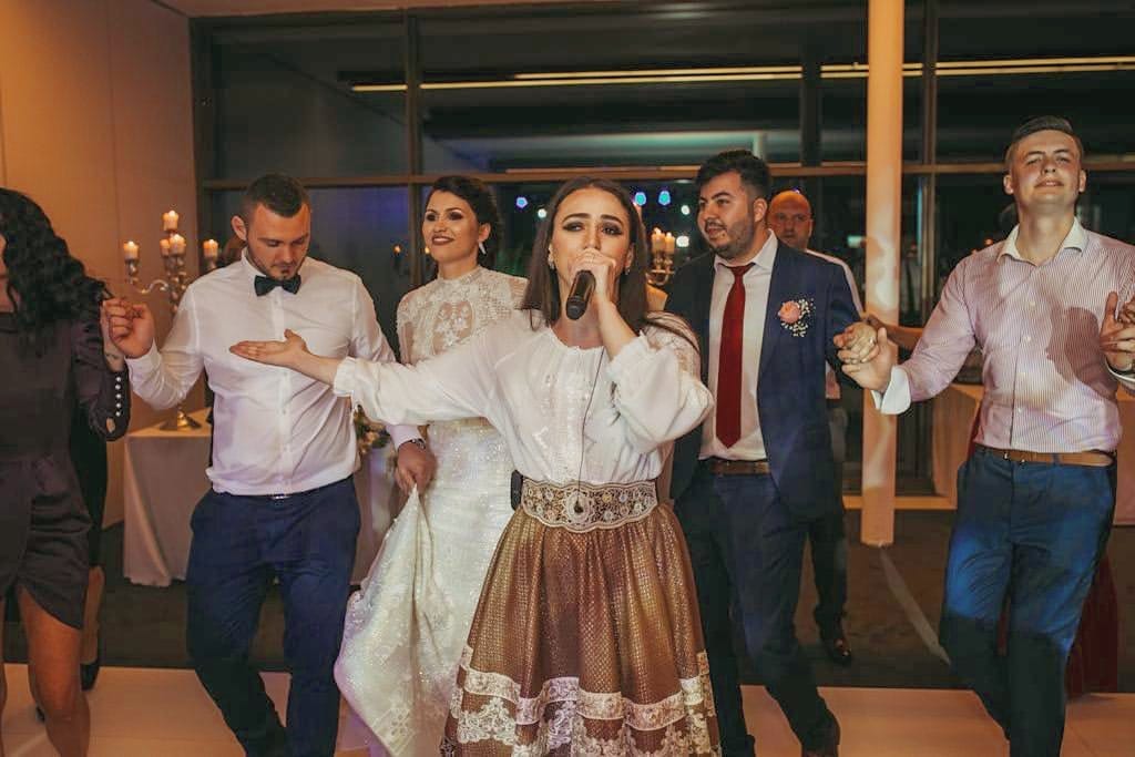 Ioana Balan cântând la microfon într-o horă de nuntă, în ie și fustă de brocart, prinsă în horă între miri și invitați