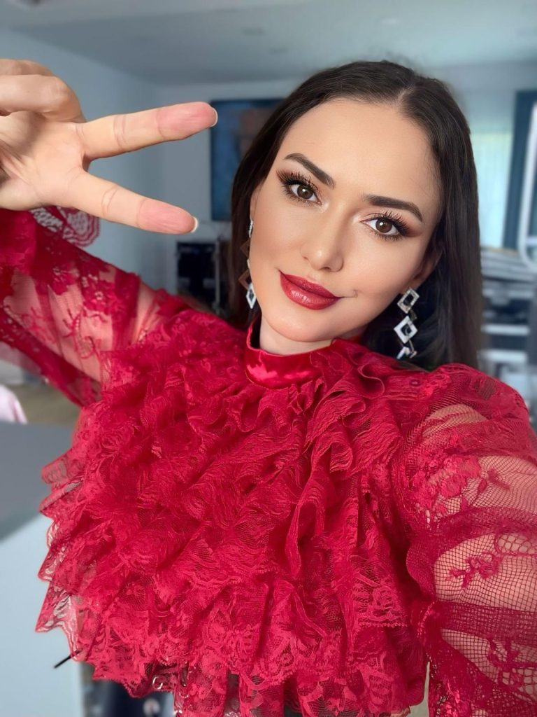 Ioana Balan în bluză roșie din dantelă cu volane, cu mâna ridicată spre obiectiv în semnul V