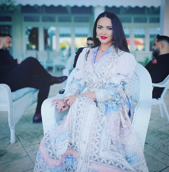 Ioana Balan așezată, în rochie albă cu imprimeu, la un eveniment în aer liber