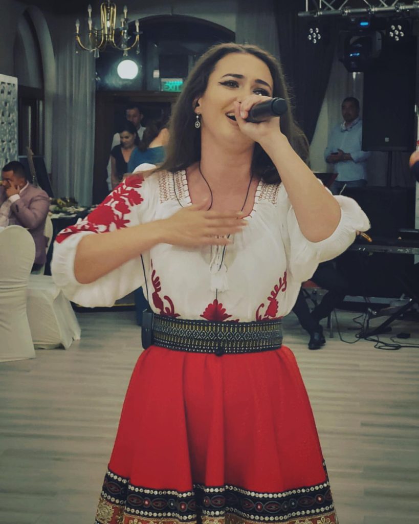 Ioana Balan cântând la microfon pe scenă, în ie cu broderie roșie și fustă roșie cu bordură neagră